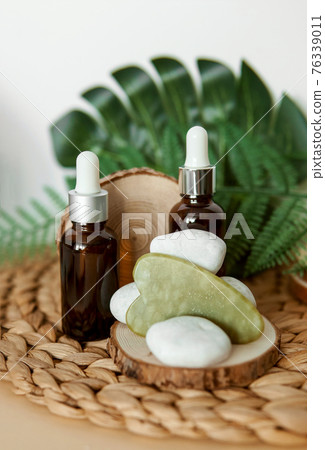 Jade Gua sha scraper and bottles of cosmetic serum for the face on wooden stands. 76339011