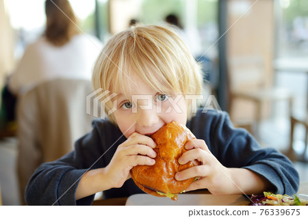 Cute preschooler boy eating large hamburger at fast food restaurant. Unhealthy meal for kids. Junk food. Overweight problem child. 76339675