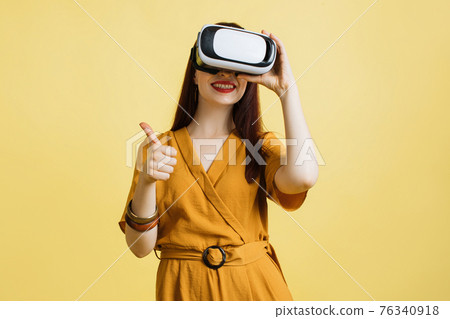 Cheerful young woman in yellow overalls, posing isolated on yellow wall background, enjoying vr, sport or games in headset, showing thumb up. People lifestyle concept. Mock up copy space. 76340918
