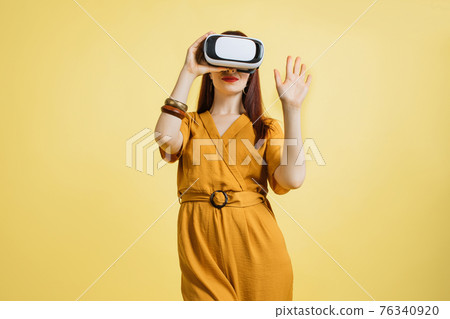 Excited young Caucasian woman in VR headset touching air exploring virtual reality at studio yellow background. Pretty lady enjoying artificial computer world or playing video game Excited young Caucasian woman in VR headset touching air exploring virtual reality at studio yellow background. Pretty lady enjoying artificial computer world or playing video game 76340920