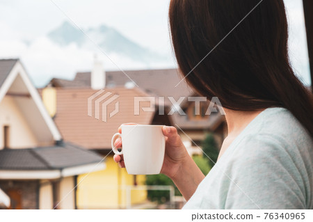 Woman standing on the balcony and drinking tea or coffee and enjoying amazing view on the mountains.  76340965