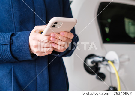 Woman using her mobile phone on the background of the charging electric car at the parking Woman using her mobile phone on the background of the charging electric car at the parking 76340967