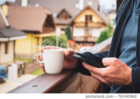 Close up man standing with a white cup and using his smartphone on the balcony on the background of modern houses. Close up man standing with a white cup and using his smartphone on the balcony on the background of modern houses. 76340968