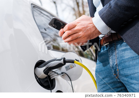 Close up a man in the suit standing near the charging electric car and using his smartphone for paying Close up a man in the suit standing near the charging electric car and using his smartphone for paying 76340971