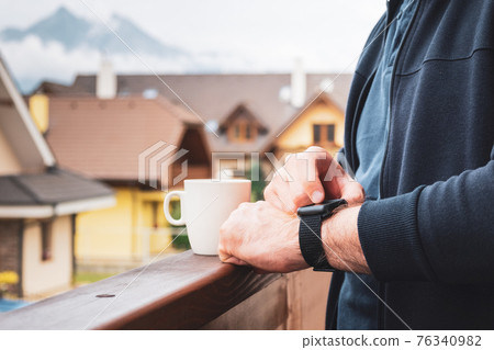 Man using his smart watch on the balcony and drinking coffee with an amazing view on the mountains and roofs of houses Man using his smart watch on the balcony and drinking coffee with an amazing view on the mountains and roofs of houses 76340982