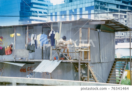 A woman reads her newspaper in a slum poor people's construciton next to luxurious buildings in Bangkok, Thailand. Retro capture on film demonstrating social inequality. A woman reads her newspaper in a slum poor people's construciton next to luxurious buildings in Bangkok, Thailand. Retro capture on film demonstrating social inequality. 76341464