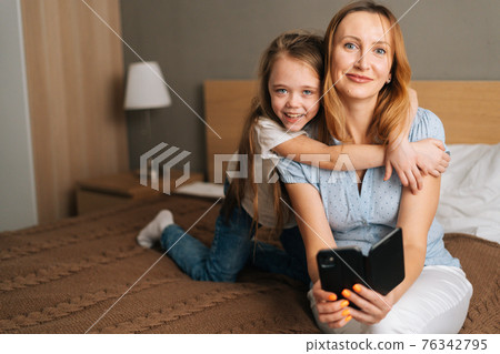 Portrait of cheerful little girl hugging to beautiful mother holding mobile phone and looking at camera, chatting on video call Portrait of cheerful little girl hugging to beautiful mother holding mobile phone and looking at camera, chatting on video call 76342795