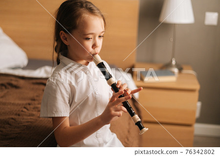 Side view of talented adorable little girl playing flute sitting on bed in bedroom, looking away. Side view of talented adorable little girl playing flute sitting on bed in bedroom, looking away. 76342802