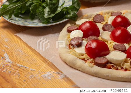 Raw pizza on baker paper on the wooden table. Preparing homemade uncooked cheese pizza with tomato sauce, tomato cherry, sausage and dough. Raw pizza on baker paper on the wooden table. Preparing homemade uncooked cheese pizza with tomato sauce, tomato cherry, sausage and dough. 76349481