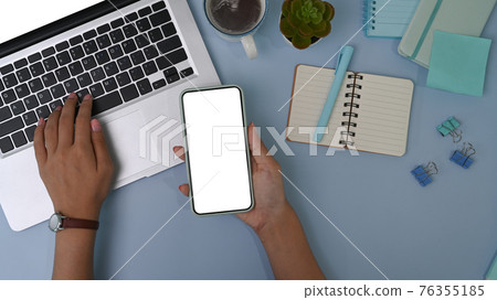 Overhead shot of woman holding smart phone and working on laptop computer on pastel blue background. 76355185