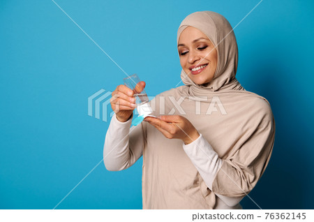 Smiling muslim woman wearing beige hijab applying micellar makeup lotion to cotton pad. Blue background, copy space 76362145