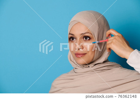 Attractive young Muslim woman with perfect makeup and clean healthy skin, posing with a syringe near her face over blue background Attractive young Muslim woman with perfect makeup and clean healthy skin, posing with a syringe near her face over blue background 76362148