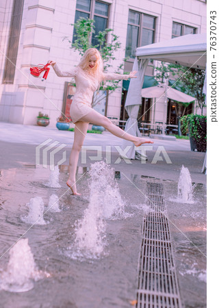 Beautiful young blonde smiling happy woman with red lips in red shoes in a white dress play near a fountain in a summer day Beautiful young blonde smiling happy woman with red lips in red shoes in a white dress play near a fountain in a summer day 76370743