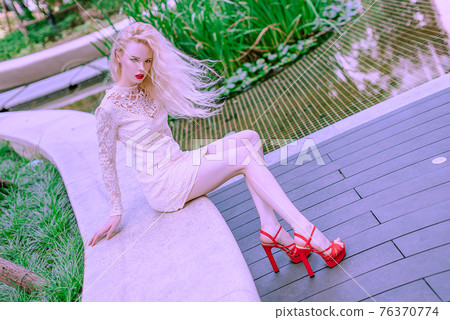 Beautiful young blonde smiling happy woman with red lips in red shoes in a white dress play near a fountain in a summer day 76370774