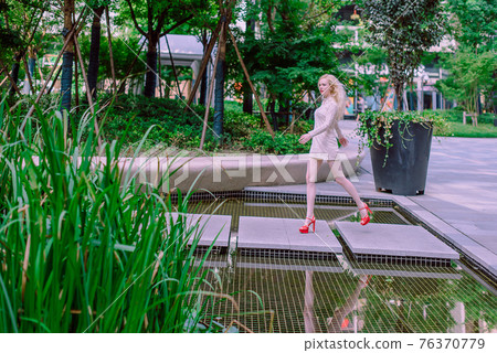 Beautiful young blonde smiling happy woman with red lips in red shoes in a white dress play near a fountain in a summer day 76370779