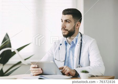 A male doctor of oriental appearance is concentratedly working on a tablet, sitting at a table in his office. 76372816
