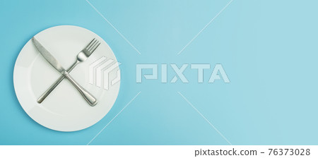 Empty plate on a blue minimal banner background. Empty white ceramic plate with knife and fork on the table after eating. Diet and healthy food concept. 76373028