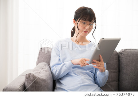 Woman with glasses sitting on the sofa and looking at the tablet device Woman with glasses sitting on the sofa and looking at the tablet device 76374073