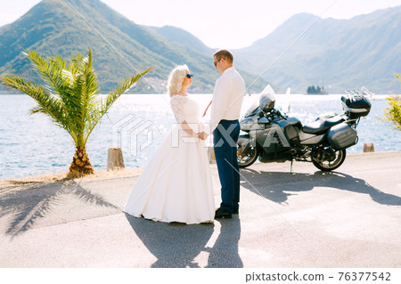 The bride and groom stand on the pier near Perast holding hands, next to them a motorcycle The bride and groom stand on the pier near Perast holding hands, next to them a motorcycle 76377542