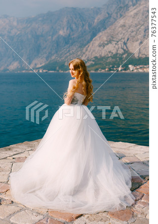 A beautiful blonde bride in a puffy white dress stands on a pier in the Bay of Kotor A beautiful blonde bride in a puffy white dress stands on a pier in the Bay of Kotor 76377543