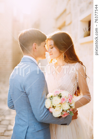 The bride and groom hug on a beautiful old street of Perast near a white stone wall, close-up The bride and groom hug on a beautiful old street of Perast near a white stone wall, close-up 76377544
