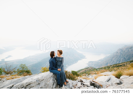 The bride and groom sitting embracing on the Lovcen mountain behind them opens a view of the Bay of Kotor The bride and groom sitting embracing on the Lovcen mountain behind them opens a view of the Bay of Kotor 76377545