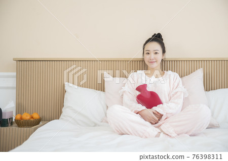 Asian woman with hair bun in pink pajamas on bed hugging rubber warm water bag Asian woman with hair bun in pink pajamas on bed hugging rubber warm water bag 76398311