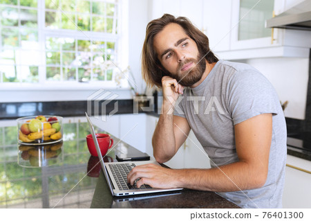 Caucasian man sitting in kitchen using laptop and thinking Caucasian man sitting in kitchen using laptop and thinking 76401300