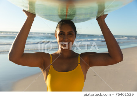 Happy mixed race woman on the beach carrying surfboard on head Happy mixed race woman on the beach carrying surfboard on head 76401529