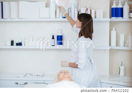 Cosmetologist in a white coat taking beauty products from the shelf Cosmetologist in a white coat taking beauty products from the shelf 76412102