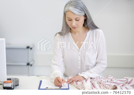 Serious woman writing information in document behind counter Serious woman writing information in document behind counter 76412570