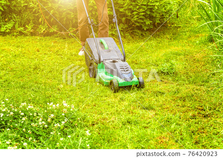 Man cutting green grass with lawn mower in backyard. Gardening country lifestyle background. Beautiful view on fresh green grass lawn in sunlight, garden landscape in spring or summer season. 76420923