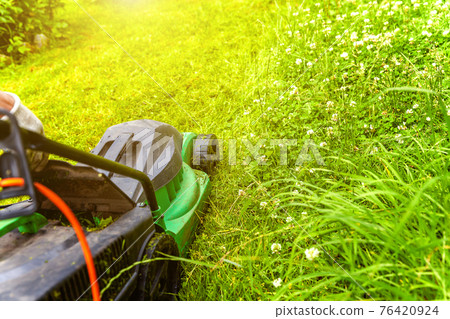 Man cutting green grass with lawn mower in backyard. Gardening country lifestyle background. Beautiful view on fresh green grass lawn in sunlight, garden landscape in spring or summer season. 76420924