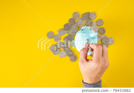 Hand puts a coin into the piggy bank coin on a yellow background. Top view Hand puts a coin into the piggy bank coin on a yellow background. Top view 76431662