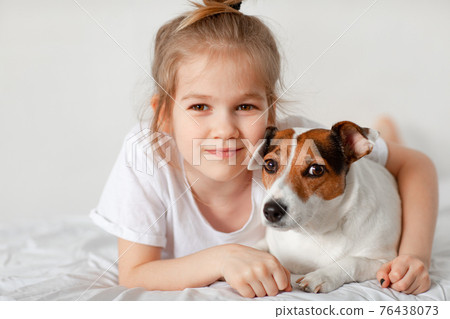 Beautiful funny kid girl lies in bed with a dog in a white bedroom. Beautiful funny kid girl lies in bed with a dog in a white bedroom. 76438073