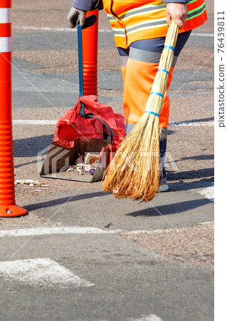 Woman janitor collects garbage from the sidewalk on a sunny day. Woman janitor collects garbage from the sidewalk on a sunny day. 76439811