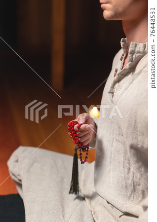 Red rosary for prayer and practice in male hands in a dark room by candlelight 76448531