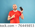 A bald middle-aged man with a beard and in a red T-shirt on a blue background. Holds a phone in his hand and points at it with the finger of his other hand 76449853