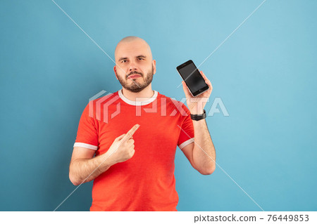 A bald middle-aged man with a beard and in a red T-shirt on a blue background. Holds a phone in his hand and points at it with the finger of his other hand A bald middle-aged man with a beard and in a red T-shirt on a blue background. Holds a phone in his hand and points at it with the finger of his other hand 76449853