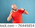 A bald middle-aged man with a beard and in a red T-shirt on a blue background. Holds a phone in his hand and points at it with the finger of his other hand 76449854