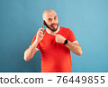 A bald middle-aged man with a beard and in a red T-shirt on a blue background. Holds a phone in his hand and points at it with the finger of his other hand 76449855