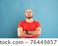 A middle-aged man with a beard in a red T-shirt on a blue background stands with his arms outstretched and rejoices in victory. Isolated. 76449857
