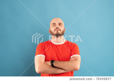 A middle-aged man with a beard in a red T-shirt on a blue background stands with his arms outstretched and rejoices in victory. Isolated. 76449857