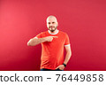 A middle-aged man with a beard in a red T-shirt on a red background stands rejoicing in victory. Isolated. 76449858