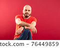 A middle-aged man with a beard in a red T-shirt on a red background joyfully points to the right. Isolated. 76449859