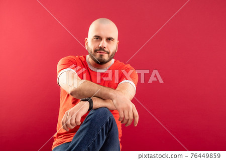 A middle-aged man with a beard in a red T-shirt on a red background joyfully points to the right. Isolated. 76449859