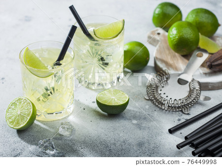 Gimlet Kamikaze cocktail in crystal glasses with lime slice and ice on light board with fresh limes and strainer. Gimlet Kamikaze cocktail in crystal glasses with lime slice and ice on light board with fresh limes and strainer. 76449998