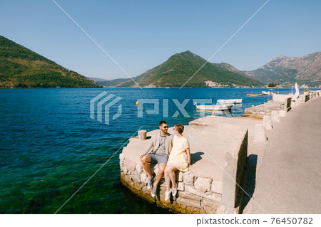 A man and a woman sit side by side on the edge of a pier near Perast in the Bay of Kotor  76450782