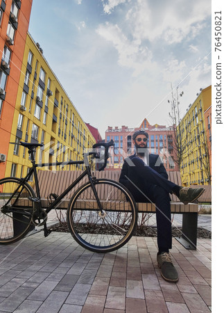 A handsome young caucasian man sits on a bench next to his bike A handsome young caucasian man sits on a bench next to his bike 76450821
