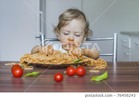Funny baby child getting messy eating spaghetti with tomato sauce from a large plate, by itself with his hands, at home Funny baby child getting messy eating spaghetti with tomato sauce from a large plate, by itself with his hands, at home 76456243